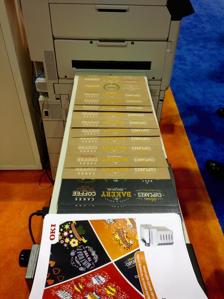 Long array of gold-foil printed sheets feeding out of a digital finishing machine at a trade show.