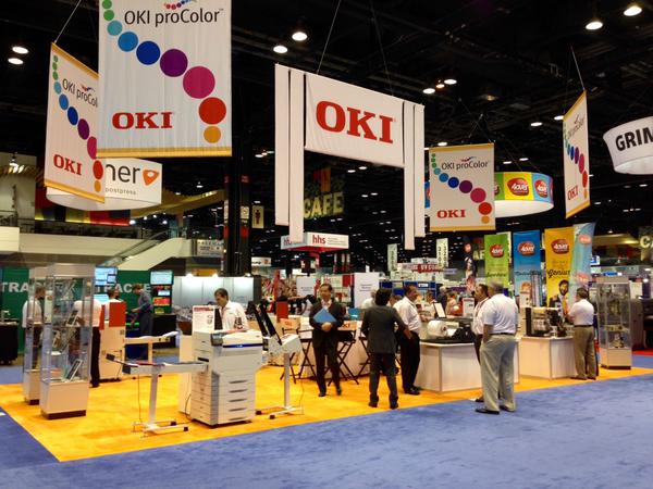 Wide view of an OKI exhibitor booth at a print expo with printers and people walking the show floor.