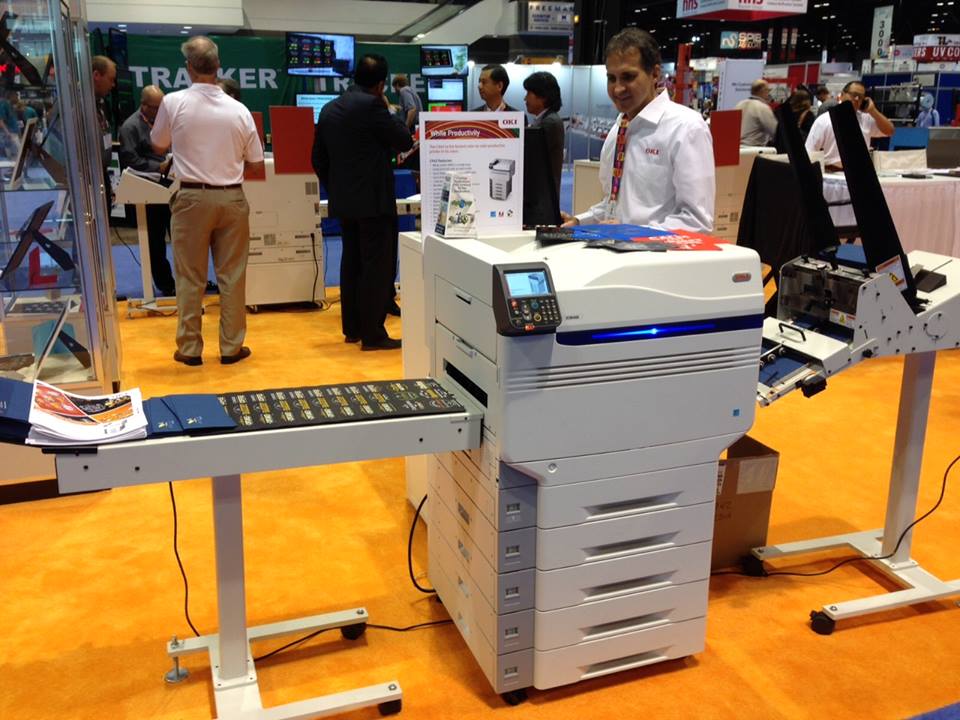 Sales representative standing beside a production color printer on an orange trade show floor.