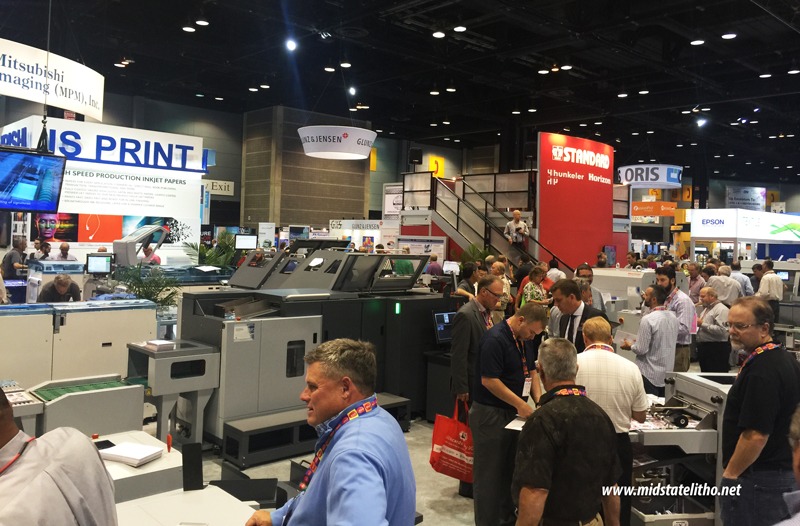 Attendees gathered around a digital print and finishing equipment setup at a busy trade show.