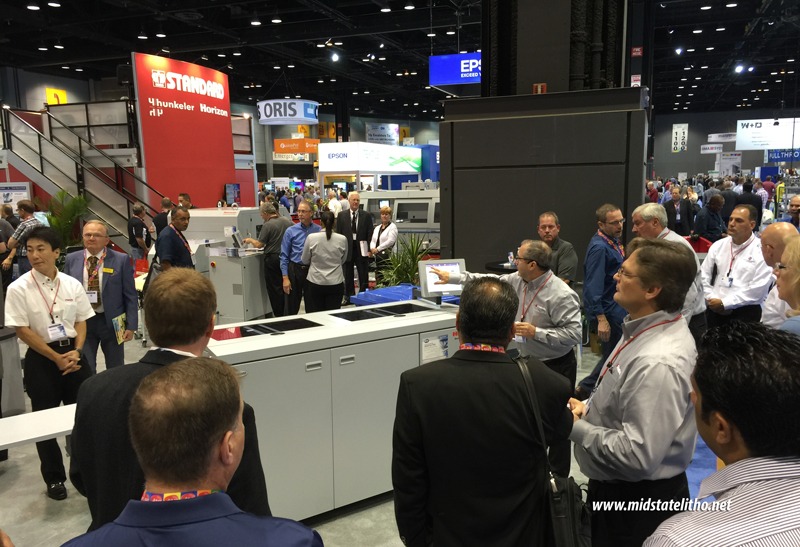 Crowded print industry trade show booth with large finishing equipment and attendees watching a live demonstration.